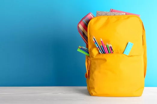 A photo of a yellow backpack stuffed with pencils, a ruler, pencil cases, and books in front of a blue background.