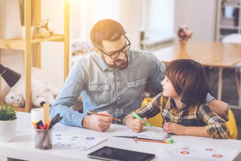 Photograph of a dad helping his child with homework at a table in their home. They are writing in a notebook and smiling at each other. School supplies and a tablet are nearby.