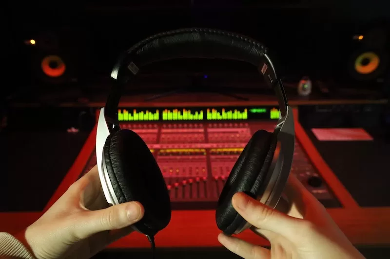 Photograph of a figure holding a pair of headphones in front of a music production work station.