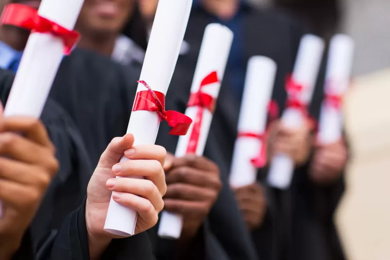 Photograph showing a row of out-of-focus graduates with their diplomas in focus in the foreground.