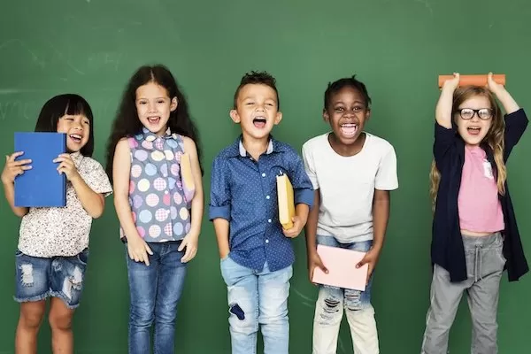 Photograph of elementary students gathered in front of a chalkboard, holding books and laughing or smiling.