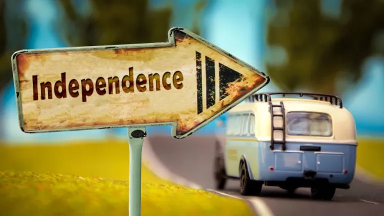 Image depicting a miniature RV travel down a road with a road sign that reads "independence" pointing to the right.