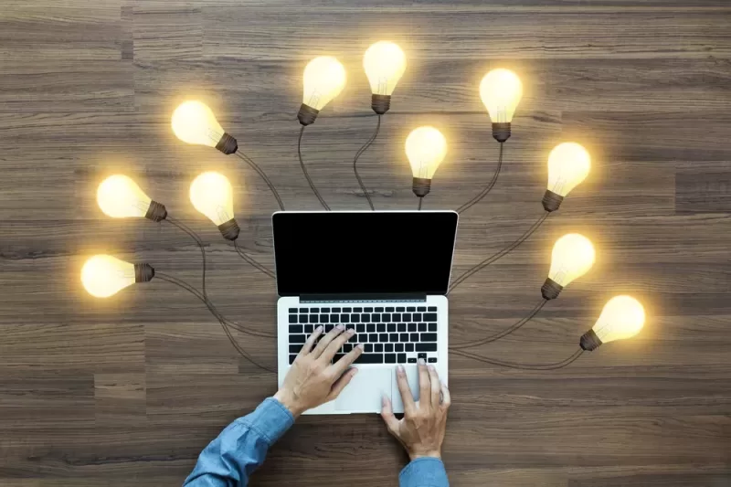 Image of a laptop on a wood-paneled surface with a collection of lightbulbs surrounding the laptop connected by wires, symbolizing ideas and inspiration. A user types on the laptop.