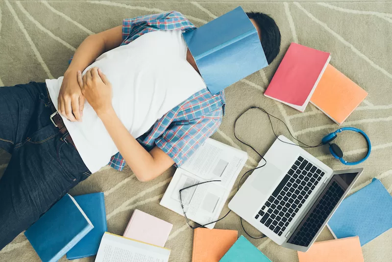 Photograph depicting a student laying beside their laptop, with an open text book, notebooks, and their glasses nearby. One book covers the students face, symbolizing the challenges associated with academic expectations.