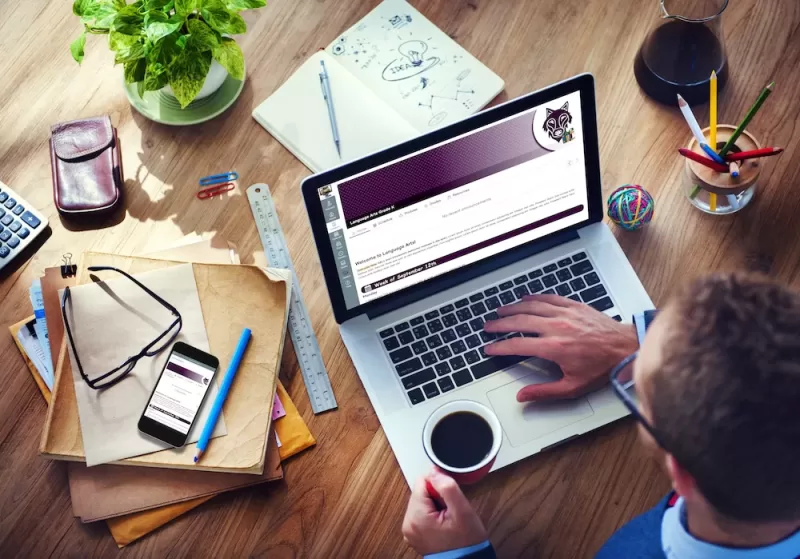 A user accesses VTVLC's Canvas instance on a laptop while working at a table which houses coffee, a notebook, pencils, and other office supplies.