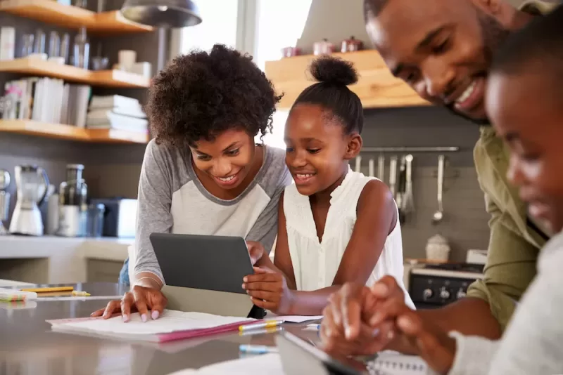 Parents work with their children on tablets at the kitchen table, which contains notebooks, pencils, and books.