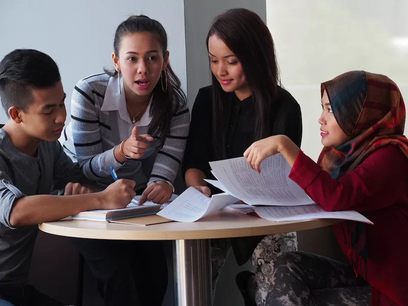 Image depicting students gathered around a table write in notebooks and engage in a discussion.