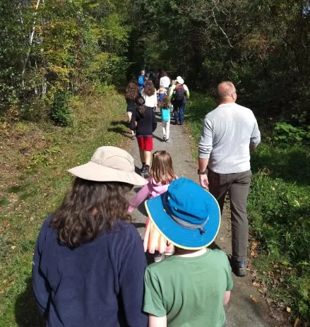 A group of students hiking on a forest trail, surrounded by tall trees and dense greenery. The students are wearing backpacks and outdoor gear, walking together in a line.