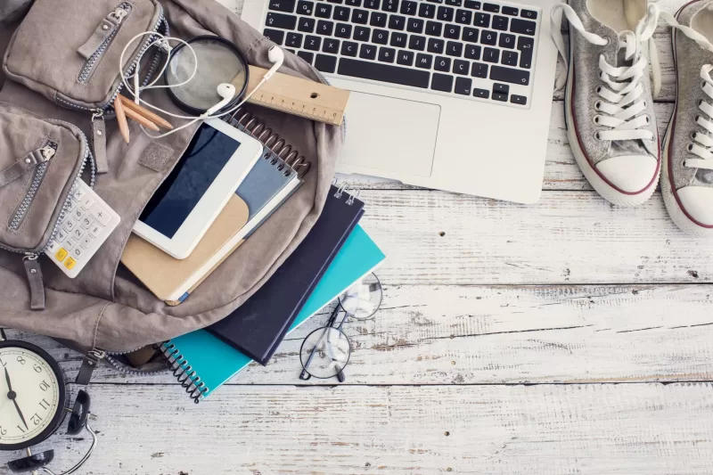 Image showing a laptop, a pair of sneakers, an alarm clock, and a backpack stuffed with various school supplies, such as pencils, a calculator, a planner, a ruler, a tablet, and headphones, arranged on a wood paneled surface.