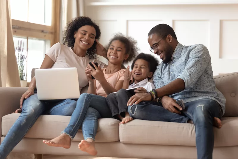 Cheerful parents and kids laugh and use devices together on sofa