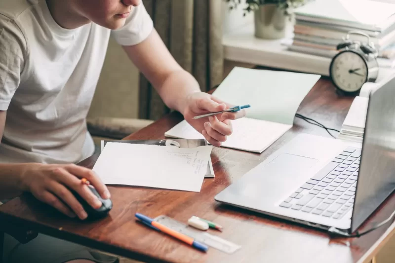 Image depicting a student writing on a piece of paper at a desk in front of a laptop.