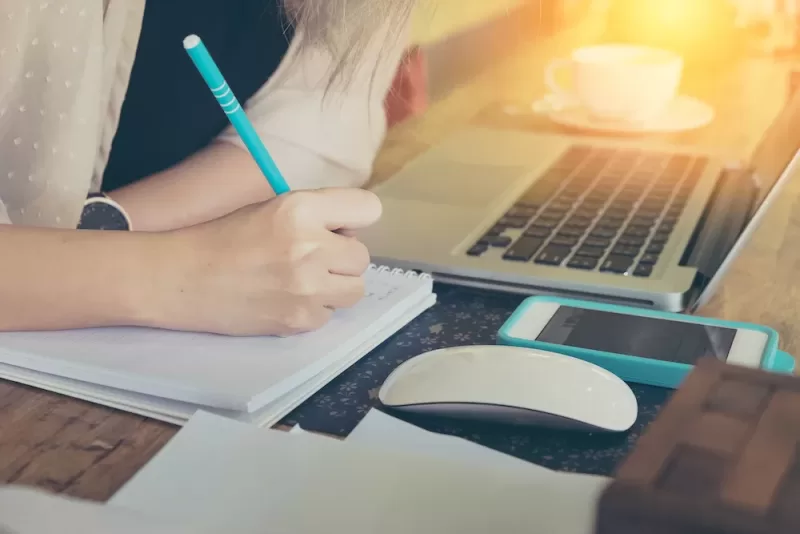 Image depicting a woman working at a desk, writing in a notebook with a laptop and computer mouse nearby.