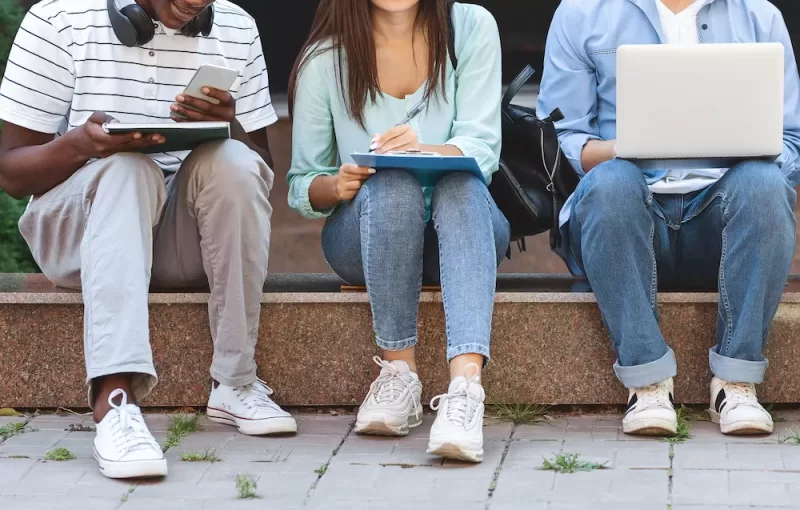 Image depicting three students working outside a school building, with notebooks, calculators, and laptops.