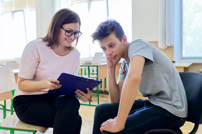 Image depicting a teacher supporting a student in a classroom. The teacher and student are looking over a clipboard.