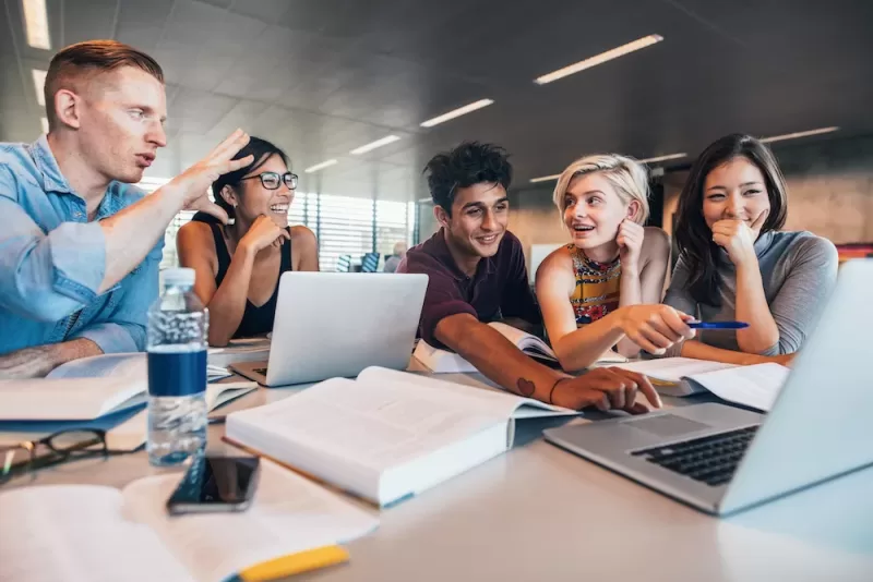 Image depicting professionals collaborating and sharing a discussion at a table with books and a laptop.