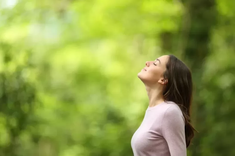 A woman stands outdoors, smiling with her arms raised in a carefree manner, enjoying a peaceful natural setting. The background is a blurred scene of green trees, possibly in a park or forest, on a sunny day. She is casually dressed, and her expression and posture suggest joy and freedom.